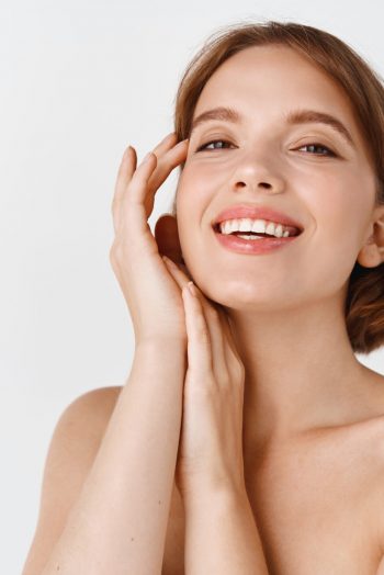 Skin care beauty. Smiling natural woman with naked shoulders and healthy clean and fresh skin, looking happy, touching cheek. Girl apply facial cosmetics, white background.